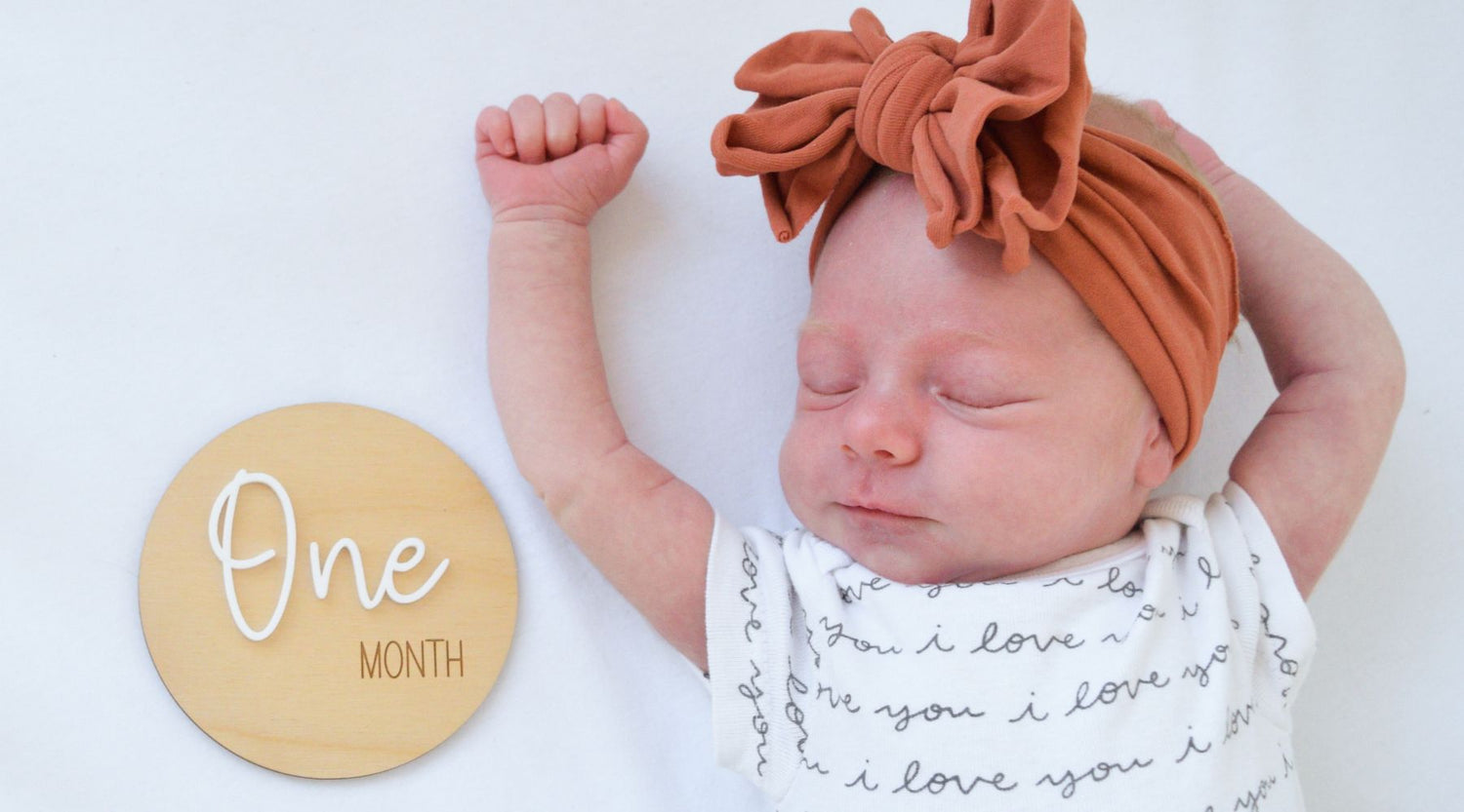 sleeping baby with arms above head laying next to a wood sign that says One Month
